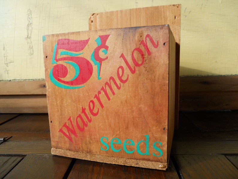 May include: A wooden box with the text "5&cent; Watermelon Seeds" printed on it in red and teal lettering.