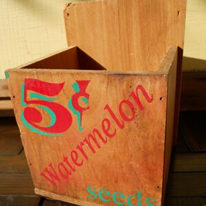 May include: A wooden box with a red and green painted design. The box says "5&cent; Watermelon Seeds".