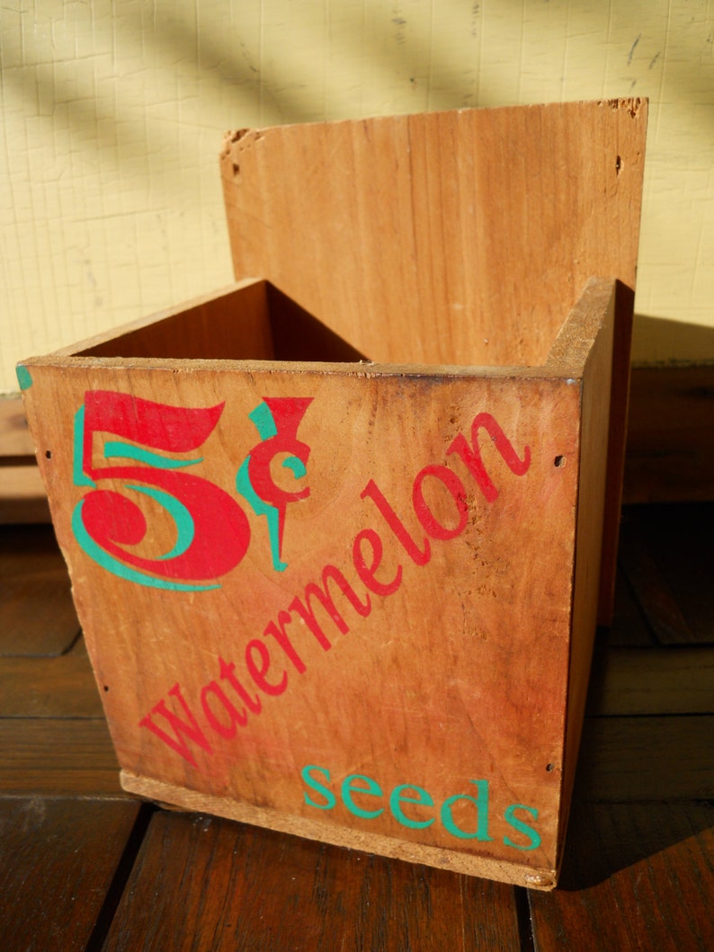 May include: A wooden box with a red and green painted design. The box says "5&cent; Watermelon Seeds".