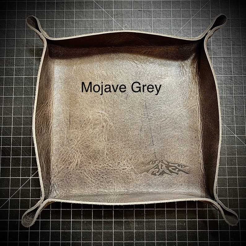 Puede incluir: Una bandeja cuadrada de cuero marr&oacute;n con esquinas redondeadas. La bandeja tiene un dise&ntilde;o decorativo en la parte inferior y el texto "Mojave Grey" est&aacute; impreso en el centro.