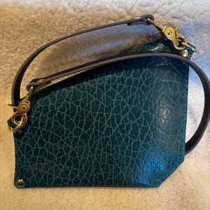May include: Teal leather crossbody bag with a textured surface and a dark gray strap. The bag features gold-colored hardware, including clasps and rivets. The strap is attached to the bag with gold-colored clasps.