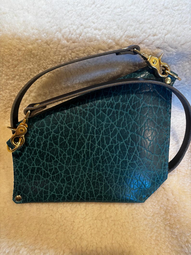 May include: Teal leather crossbody bag with a textured surface and a dark gray strap. The bag features gold-colored hardware, including clasps and rivets. The strap is attached to the bag with gold-colored clasps.