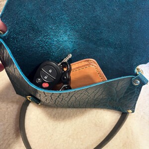 May include: A teal leather crossbody bag with a black strap. Inside the open bag are car keys and a small brown leather wallet. The bag has a textured surface and gold-colored hardware.