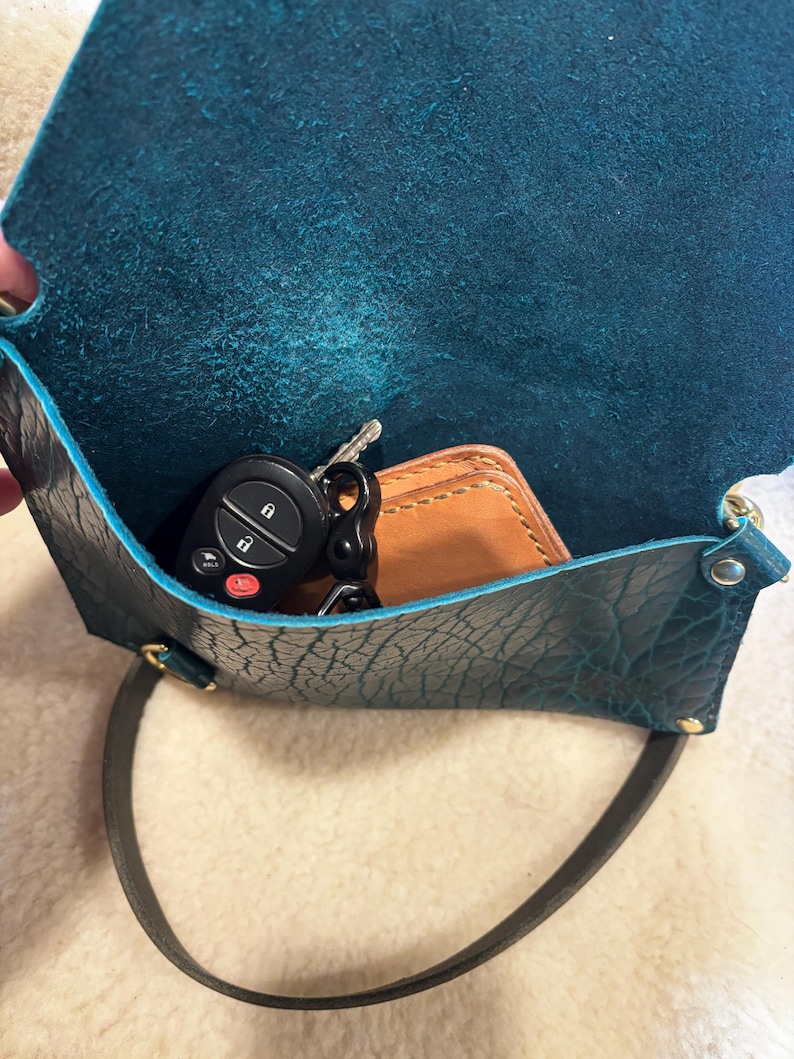 May include: A teal leather crossbody bag with a black strap. Inside the open bag are car keys and a small brown leather wallet. The bag has a textured surface and gold-colored hardware.