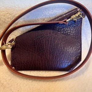 May include: A dark burgundy leather crossbody bag with a textured surface. The bag has a brown leather strap and gold-colored metal hardware. The strap is adjustable and can be worn over the shoulder or across the body.
