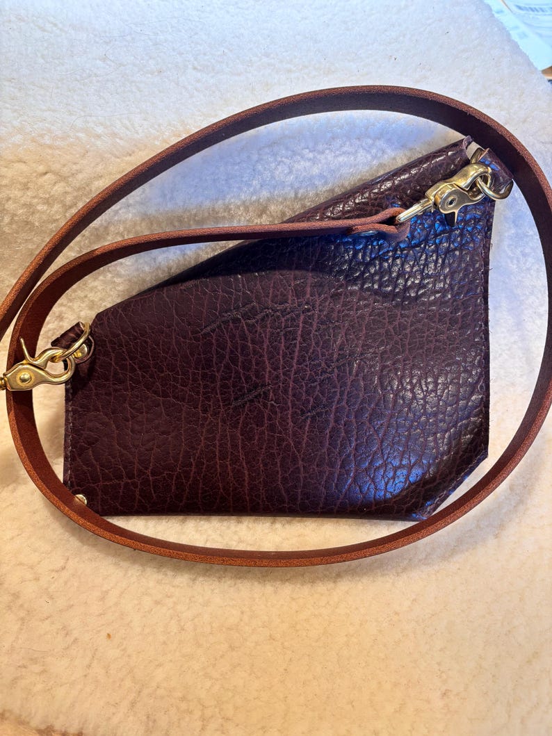 May include: A dark burgundy leather crossbody bag with a textured surface. The bag has a brown leather strap and gold-colored metal hardware. The strap is adjustable and can be worn over the shoulder or across the body.