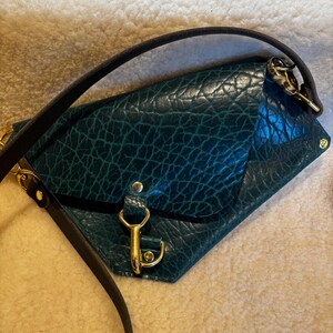 May include: A teal leather crossbody bag with a textured surface. The bag features gold-colored hardware, including a clasp and strap attachments. A black strap is attached to the bag.