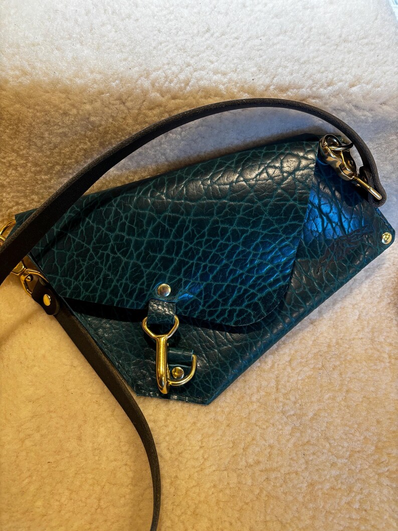 May include: A teal leather crossbody bag with a textured surface. The bag features gold-colored hardware, including a clasp and strap attachments. A black strap is attached to the bag.