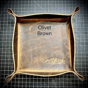 Puede incluir: Una bandeja de cuero marr&oacute;n con un dise&ntilde;o de monta&ntilde;a en relieve en la parte inferior. La bandeja tiene un borde doblado y est&aacute; etiquetada como "Olivet Brown".