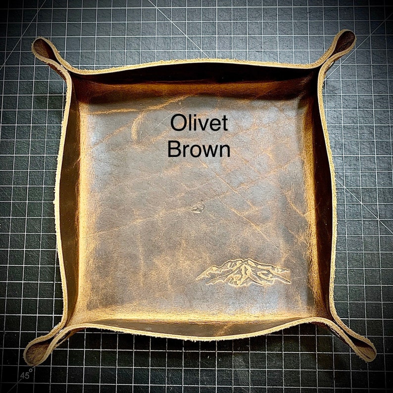 Puede incluir: Una bandeja de cuero marr&oacute;n con un dise&ntilde;o de monta&ntilde;a en relieve en la parte inferior. La bandeja tiene un borde doblado y est&aacute; etiquetada como "Olivet Brown".