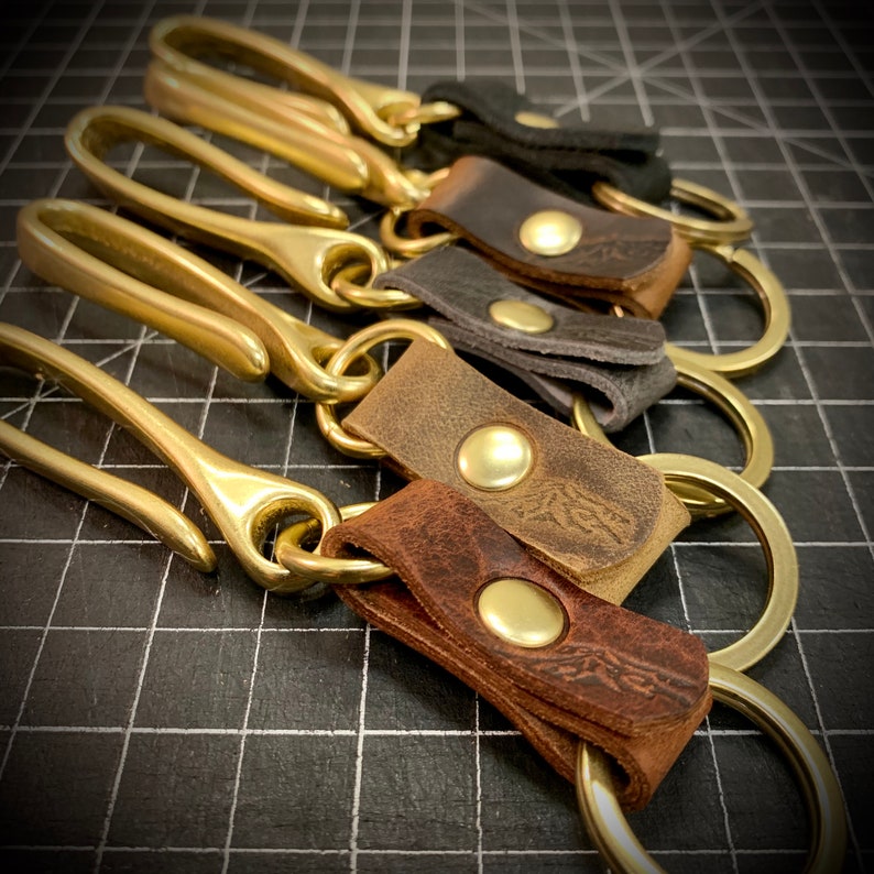Op de afbeelding: Een set van vijf messing sleutelhangers met leren lussen. Elke sleutelhanger heeft een leren lus in een andere kleur, waaronder bruin, beige, grijs en zwart.