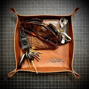 Puede incluir: Una bandeja de cuero marr&oacute;n con un dise&ntilde;o de monta&ntilde;a. La bandeja contiene un par de gafas de sol, una cartera, un juego de llaves y un par de auriculares.