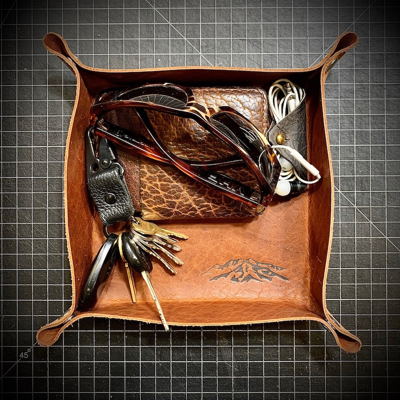 Puede incluir: Una bandeja de cuero marr&oacute;n con un dise&ntilde;o de monta&ntilde;a. La bandeja contiene un par de gafas de sol, una cartera, un juego de llaves y un par de auriculares.