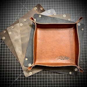 May include: A brown leather tray with a mountain design, surrounded by four other leather trays in various shades of brown and gray.