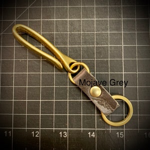 Op de afbeelding: Sleutelhanger van messing met een leren lus en een messing ring. De leren lus is gegraveerd met de tekst "Mojave Grey".