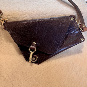 May include: A dark brown leather crossbody bag with a textured surface. The bag has a flap closure with a gold-colored clasp and a long, thin leather strap. Gold-colored hardware accents the bag's corners.