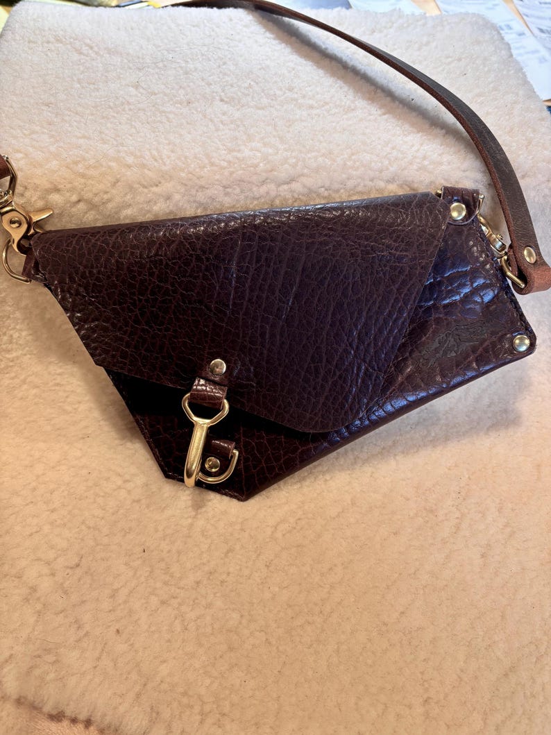 May include: A dark brown leather crossbody bag with a textured surface. The bag has a flap closure with a gold-colored clasp and a long, thin leather strap. Gold-colored hardware accents the bag's corners.
