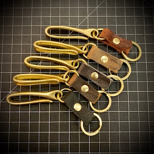 Op de afbeelding: Vijf messing sleutelhangers met leren lussen. De sleutelhangers zijn op elkaar gestapeld. De leren lussen zijn in verschillende kleuren, waaronder bruin, beige en zwart.