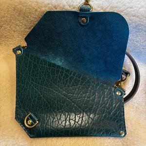 May include: Open teal leather crossbody bag with a textured exterior and a smooth interior. The bag features gold-colored hardware, including a clasp and rivets. A black strap is attached.