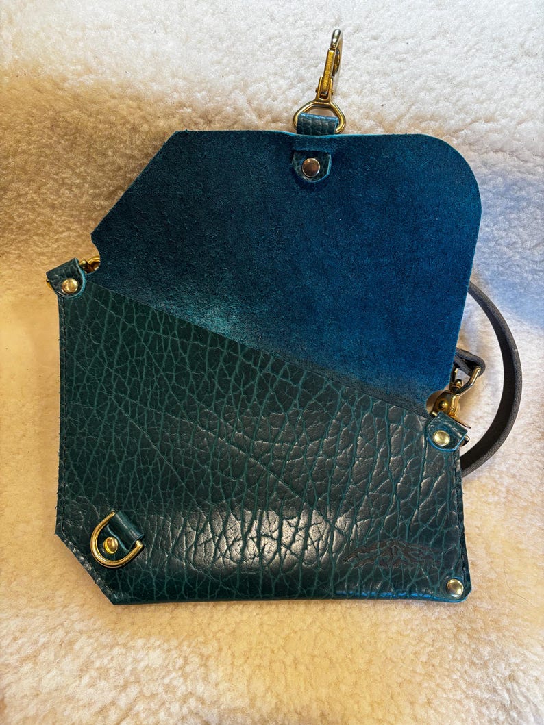 May include: Open teal leather crossbody bag with a textured exterior and a smooth interior. The bag features gold-colored hardware, including a clasp and rivets. A black strap is attached.