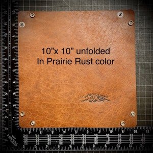 Puede incluir: Una bandeja de cuero de 25,4 cm x 25,4 cm en color marr&oacute;n llamada "Prairie Rust". La bandeja est&aacute; desplegada y tiene un dise&ntilde;o de cordillera en relieve en el cuero. La bandeja tiene cuatro broches blancos alrededor de los bordes.