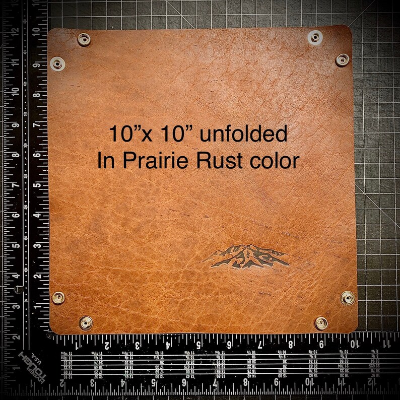 Puede incluir: Una bandeja de cuero de 25,4 cm x 25,4 cm en color marr&oacute;n llamada "Prairie Rust". La bandeja est&aacute; desplegada y tiene un dise&ntilde;o de cordillera en relieve en el cuero. La bandeja tiene cuatro broches blancos alrededor de los bordes.