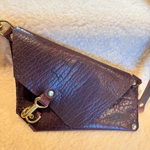 May include: A dark brown leather crossbody bag with a textured surface. The bag features a gold-colored clasp and hardware. The strap is also brown leather. The bag is designed with a unique, angular shape.