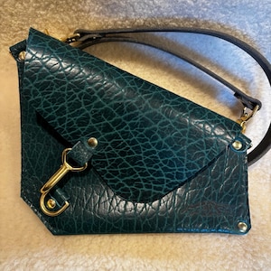 May include: A teal leather crossbody bag with a textured surface. The bag features a flap closure, gold-colored hardware, and a black strap. The bag is rectangular with a triangular flap.