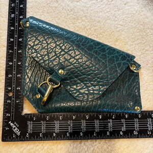 May include: Teal leather clutch with a textured finish. The clutch features a flap closure with a gold-tone clasp and hardware. The clutch measures approximately 11 inches long.