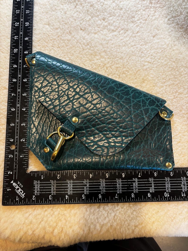 May include: Teal leather clutch with a textured finish. The clutch features a flap closure with a gold-tone clasp and hardware. The clutch measures approximately 11 inches long.