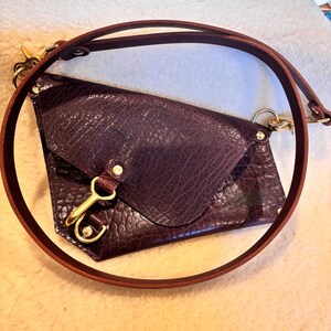 May include: A deep burgundy leather crossbody bag with a textured surface. The bag features a flap closure, a gold-colored clasp, and a matching brown leather strap. The strap is adjustable and can be worn over the shoulder or across the body.