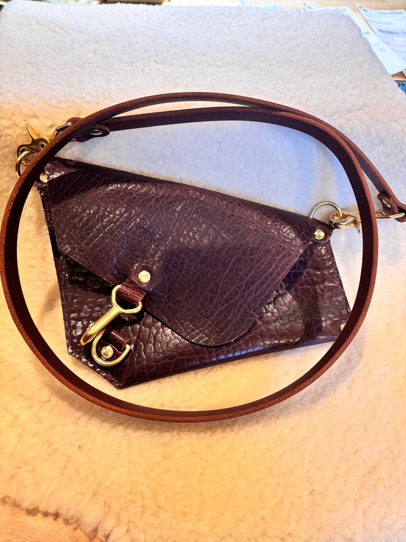 May include: A deep burgundy leather crossbody bag with a textured surface. The bag features a flap closure, a gold-colored clasp, and a matching brown leather strap. The strap is adjustable and can be worn over the shoulder or across the body.