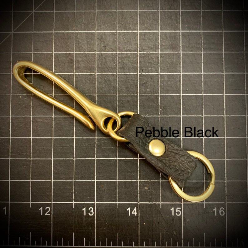 Op de afbeelding: Sleutelhanger van messing met een zwarte leren lus en een messing haak. De leren lus heeft de tekst "Pebble Black" erop gegraveerd.
