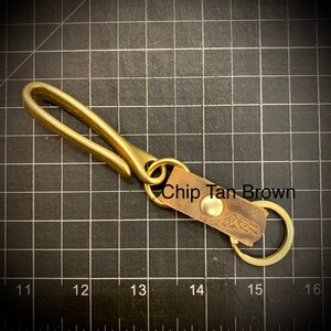 Op de afbeelding: Sleutelhanger van messing met een leren band en een messing haak. De leren band heeft de tekst "Chip Tan Brown" erop.