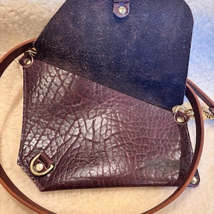 May include: A deep purple leather crossbody bag with a textured exterior and a brown leather strap. The bag has a flap closure with a gold-colored clasp and hardware. The interior is a darker shade of purple.