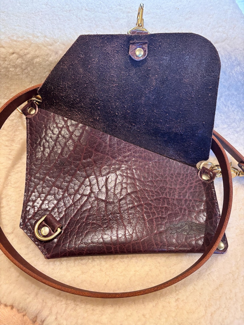 May include: A deep purple leather crossbody bag with a textured exterior and a brown leather strap. The bag has a flap closure with a gold-colored clasp and hardware. The interior is a darker shade of purple.
