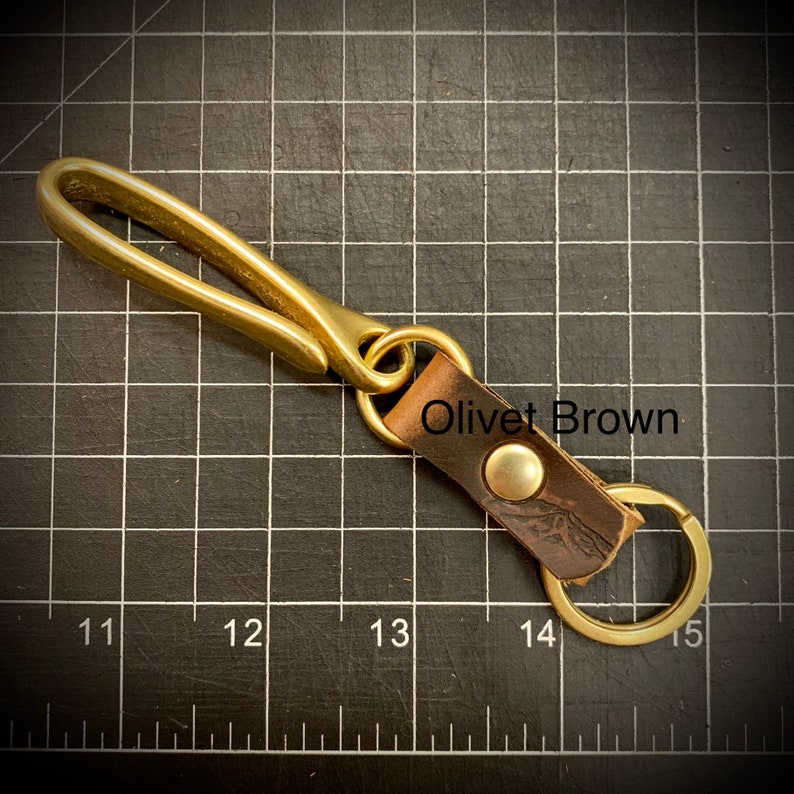 Op de afbeelding: Sleutelhanger van messing met een leren lus en een messing haak. De leren lus heeft de tekst "Olivet Brown" erop gegraveerd.