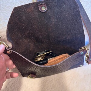May include: Open brown leather crossbody bag with a flap closure. The bag has a long, adjustable strap and gold-colored hardware. Inside, a wallet and keys are visible. The leather has a textured appearance.