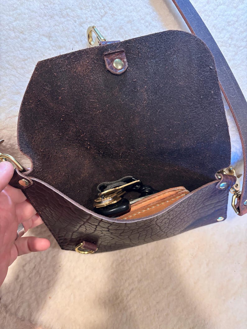May include: Open brown leather crossbody bag with a flap closure. The bag has a long, adjustable strap and gold-colored hardware. Inside, a wallet and keys are visible. The leather has a textured appearance.