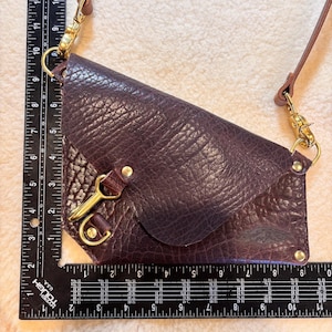 May include: A deep purple leather crossbody bag with a textured finish. The bag features a flap closure, gold-toned hardware, and a brown leather strap. The bag measures approximately 10 inches in length.