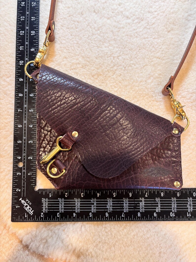 May include: A deep purple leather crossbody bag with a textured finish. The bag features a flap closure, gold-toned hardware, and a brown leather strap. The bag measures approximately 10 inches in length.