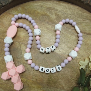 May include: A pastel-coloured rosary necklace with the names "MIA" and "ROSE" spelled out in white letter beads. The necklace features lavender, pink, and white beads, and a pink cross pendant.