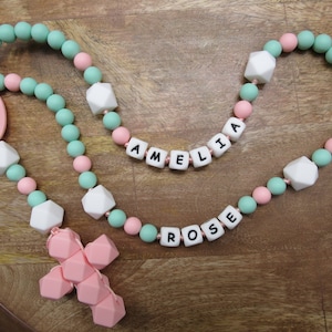 Könnte beinhalten: Eine rosa und grüne Silikon-Beißkette mit einem rosa Kreuzanhänger. Die Kette hat weiße Perlen und die Namen "Amelia" und "Rose" sind in schwarzen Buchstaben geschrieben.