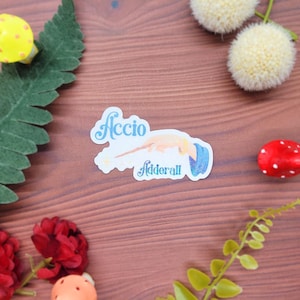 May include: A sticker with the words "Accio Adderall" in light blue script, featuring a wand and a blue scroll. The sticker is surrounded by decorative elements including red and yellow mushrooms, flowers, and greenery.