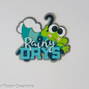 May include: A green frog with big eyes sits under a blue and green umbrella with the words "Rainy Days" in blue and black letters.