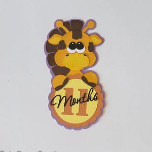May include: A brown and yellow giraffe with big eyes holds a yellow circle with the number 11 and the word "Months" written in black.