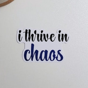 May include: White sticker with the text "i thrive in chaos" in blue cursive lettering.