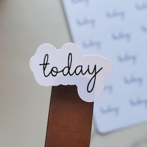 May include: A white sticker in the shape of a cloud with the word "today" in black script. A sheet of stickers with the word "today" is in the background. A small potted plant is in the upper left corner.