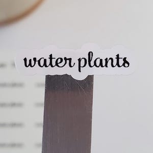 May include: A white sticker with black text that reads "water plants". The sticker is attached to a silver metal ruler.
