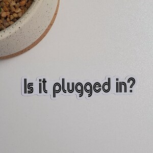 May include: White sticker with black text that reads "Is it plugged in?" on a white background with a potted cactus in the top left corner.